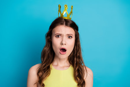 Adorable Woman in Yellow Dress with Golden Crown Against Bright Blue Background Showing Surprised Expressionの写真素材