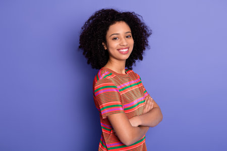 Confident young woman standing with arms crossed against a vibrant purple background wearing striped casual t-shirtの写真素材