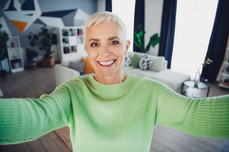 Smiling mature woman enjoying a relaxing moment at home in a vibrant green sweater, appreciating the...