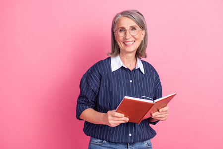 Cheerful mature woman holding a notebook against a vibrant pink background, expressing positive...