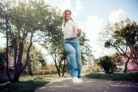Young woman enjoying a sunny day outdoors in a city park, jumping happily surrounded by greenery and...