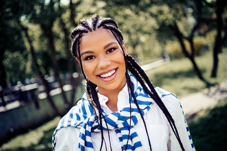 Cute beautiful woman with braided hair smiling brightly in a sunny urban park with lush greenery and...
