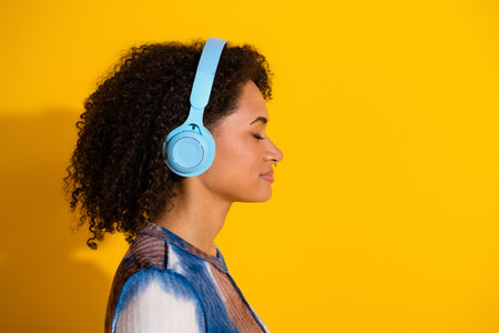 Young woman listening to music with blue headphones against a bright yellow background, expressing leisure and relaxationの写真素材