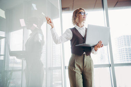 Confident businesswoman presenting ideas in a modern office holding a laptop in a bright workspaceの写真素材