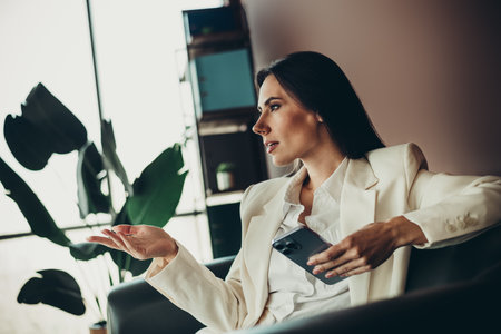 Confident businesswoman gesturing while holding a smartphone in a modern office environment during thoughtful discussionの写真素材