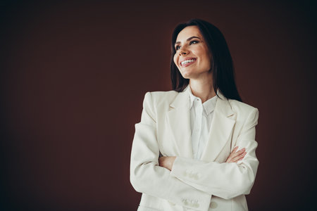 Beautiful professional businesswoman in white formalwear posing with arms crossed and smiling, brown backgroundの写真素材