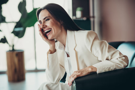Successful businesswoman in a white formal suit laughing joyfully while sitting in a modern office environment.の写真素材