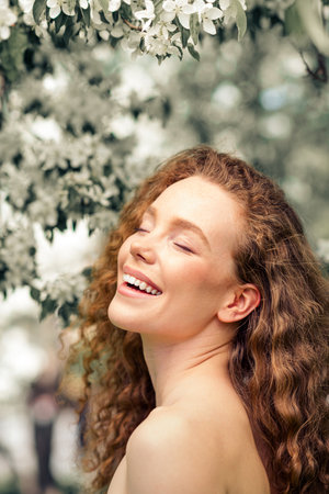 Young woman with curly red hair enjoys blossoming park in daylight, exuding joy and femininity in...