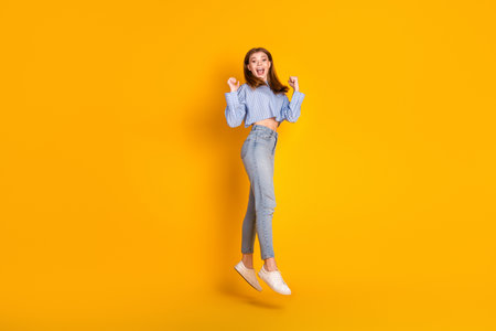 Joyful young woman jumping in excitement wearing stylish casual attire against vibrant yellow...