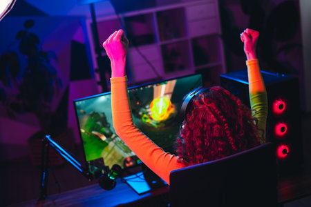 Joyful female gamer celebrates virtual gaming victory with lifted arms, enjoying an exciting evening with vibrant neon lightsの写真素材