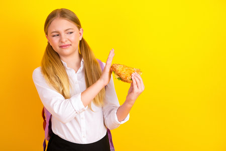 Young schoolgirl rejecting offered food item with a displeased gesture against a vibrant yellow...