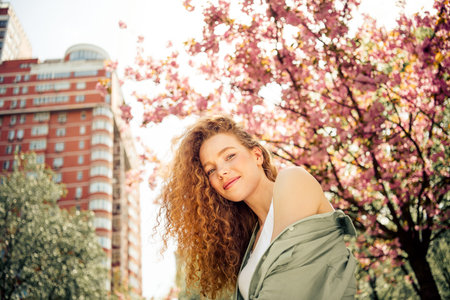 Young woman with curly red hair enjoying a sunny day outdoors among cherry blossoms in urban city...