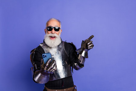 Elderly man in knight armor using smartphone while gesturing on violet backgroundの写真素材