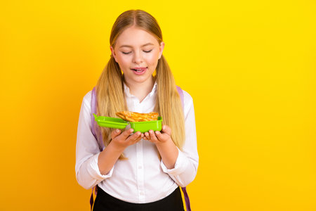 Youthful schoolgirl holding a green lunchbox with pizza against a vibrant yellow background showcasing education and snack timeの写真素材