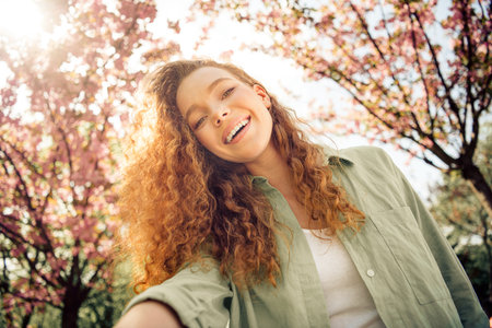 Young woman enjoying a sunny day outdoors in spring among blooming trees, smiling brightly with curly red hairの写真素材