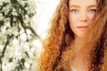 Charming young woman with curly red hair enjoying a sunny day outdoors surrounded by blooming...