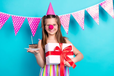 Joyful girl in colorful party outfit holding gift and cake slice with polka dot bunting on summer holiday celebrationの写真素材