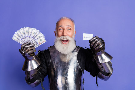 Formal knight in armor holding cash and a credit card against a purple backgroundの写真素材