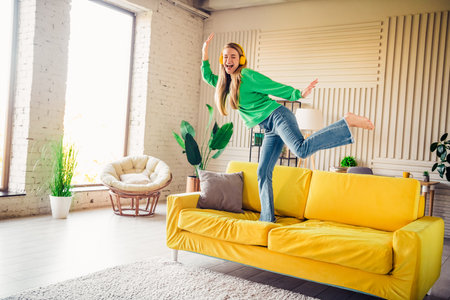 Joyful young woman enjoying music and dancing playfully on a yellow couch in a stylish, cozy loft apartmentの写真素材