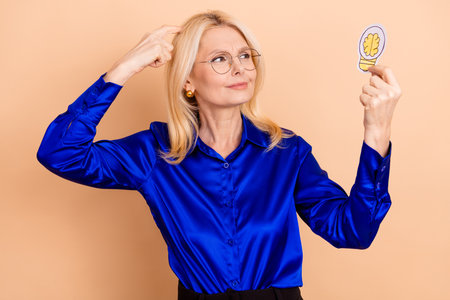 Mature woman in blue blouse holding lightbulb cartoon contemplating ideas on beige background