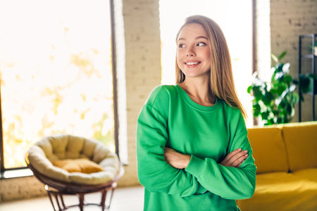 Young woman in green sweatshirt relaxing at home with crossed arms in cozy vibrant living room interiorの写真素材