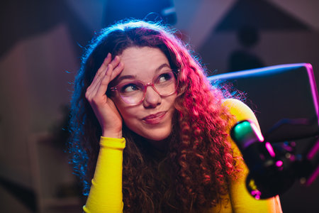 Young woman with curly hair in neon lights looking thoughtful while streaming, surrounded by vibrant colors and modern feelの写真素材
