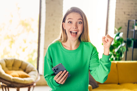 Young woman in green sweatshirt celebrating victory indoors during daylight, holding smartphone and expressing excitementの写真素材