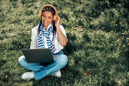 Charming woman enjoying leisure time outdoors with laptop, headphones, and smile during a sunny...