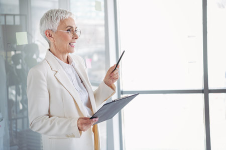Confident senior businesswoman wearing formal attire presenting a professional concept indoors using a clipboard and penの写真素材
