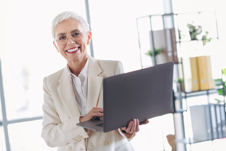 Elegant senior businesswoman in formal attire holding laptop and smiling confidently in modern office settingの写真素材