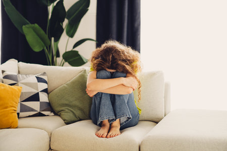 Upset woman with curly hair embracing her knees on a sofa in a cozy living room with stylish decor and natural lightの写真素材