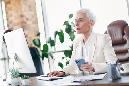 Professional businesswoman with short white hair working in a modern office using a computer and smartphoneの写真素材