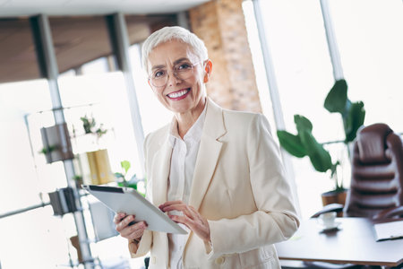 Confident senior businesswoman using a tablet in light modern office workspace, showcasing maturity and professionalismの写真素材