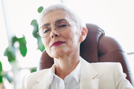 Elegant senior businesswoman relaxed in the workplace with eyes closed, exuding confidence and professionalismの写真素材