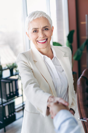 Confident senior businesswoman in formal attire greeting with a handshake in a bright modern office environmentの写真素材