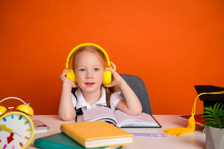 Little girl wearing headphones studies at desk with school supplies on vibrant orange backgroundの写真素材