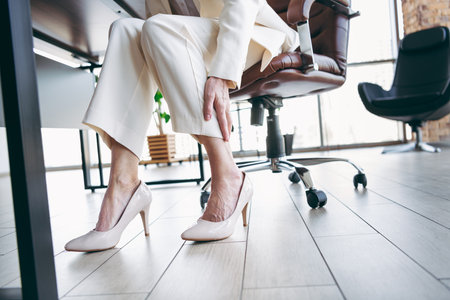 Businesswoman sitting in office adjusting high heels while working in bright and modern workplace environment.の写真素材