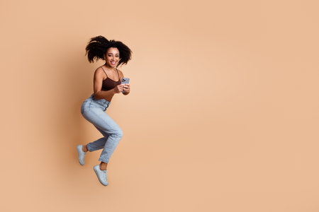 Joyful Young Woman Leaping in Stylish Clothes Holding a Smartphone Against a Beige Background