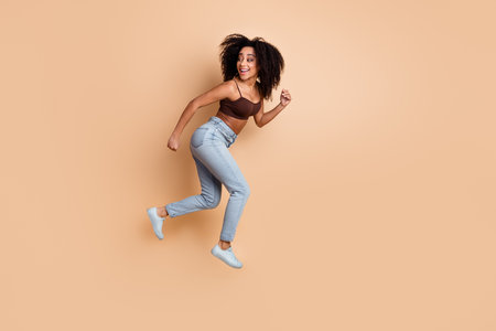 Happy young woman in casual stylish attire jumping joyfully with a vibrant hairstyle on a beige...