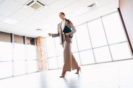 Confident businesswoman in formal attire walking in a modern office interior holding a file folder during workdayの写真素材