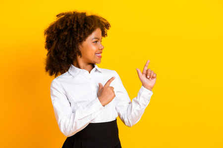 Smiling schoolgirl in a white shirt pointing with both hands against a yellow background showcasing a bright gestureの写真素材