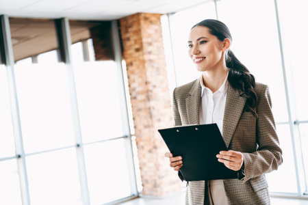 Young specialist young businesswoman holding a clipboard standing in a modern bright office interior, professional attitudeの写真素材