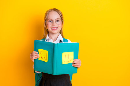 Smiling schoolgirl with glasses holding a teal notebook over yellow background, symbolizing...