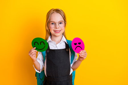 Smiling schoolgirl holding emoticon symbols expressing happy and sad emotions near a yellow...