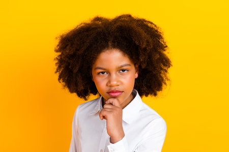 Confident schoolgirl posing with thoughtful expression against a vibrant yellow background, wearing a white formal shirtの写真素材