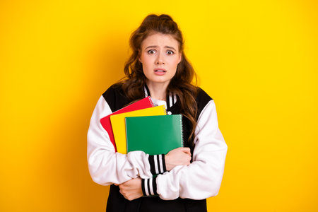 Concerned female student holding colorful notebooks on a bright yellow background wearing casual attire for schoolの写真素材