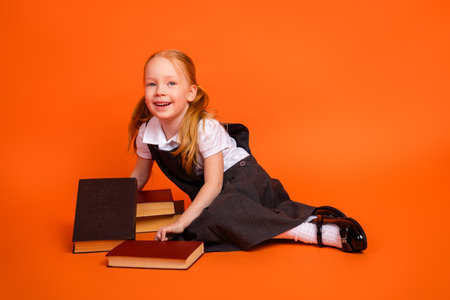 Young schoolgirl in uniform sitting with books against a bright orange background, showcasing education and learningの写真素材