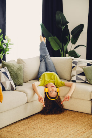 Cheerful woman in a green shirt relaxing upside down on a sofa, wearing headphones and enjoying leisure time at homeの写真素材