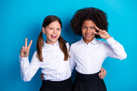 Two smiling schoolgirls in white uniforms posing with peace signs on a blue background, showing...