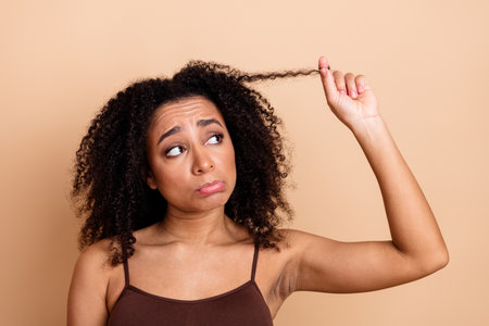 Young woman with curly black hair looks thoughtful while examining a strand against a beige backgroundの写真素材